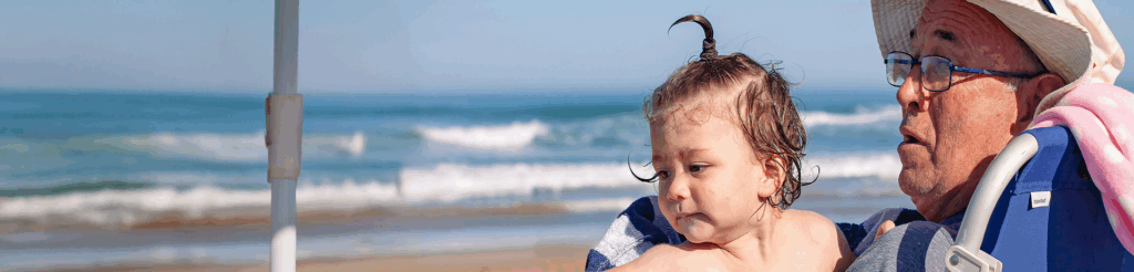 A close-up of a young child with their hair tied up in a sprout ponytail, sitting on the lap of an older man wearing a straw hat and glasses, both looking out over the ocean on a sunny beach day.