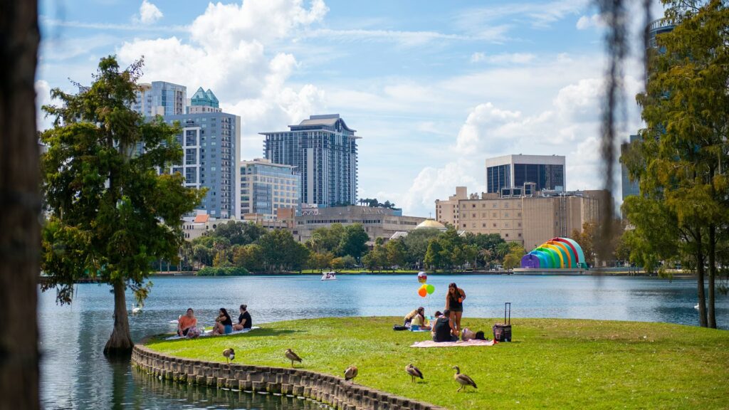 People picnic on the grassy edge of Lake Eola in downtown Orlando, with a modern city skyline and the rainbow amphitheater dome visible across the water.