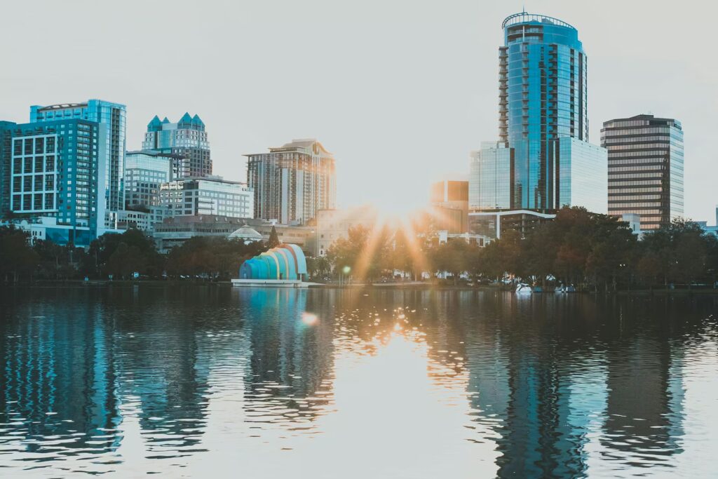 Downtown Orlando skyline reflecting in a lake, with the sun setting behind the buildings and causing a lens flare over the water.