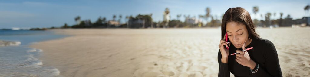 A panoramic shot of a young Black woman sitting on a wide, sunny beach, talking on a pink cell phone. She is wearing a black top and is holding a pen. The background features white sand, gentle ocean waves, and distant palm trees and buildings.