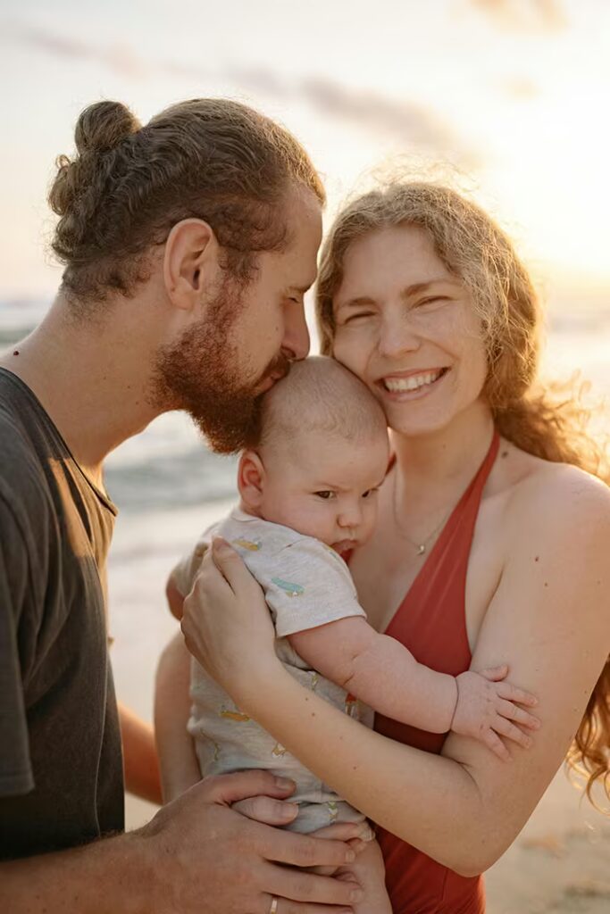 A close-up, sunlit outdoor portrait of a couple holding their baby. The man, with a beard and hair pulled up in a bun, is kissing the baby's forehead. The woman smiles directly at the camera, holding the baby close. They are standing near a beach with the ocean in the background.